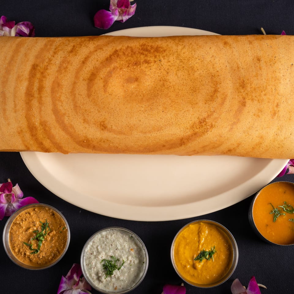 A long, golden-brown dosa on a white plate, surrounded by four small bowls of colorful chutneys.